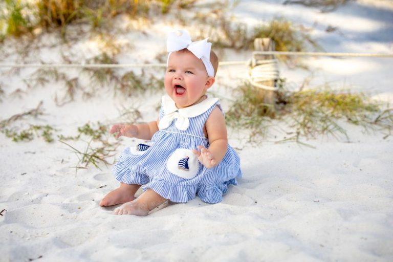 Baby in white outfit crawling and playing on sandy beach near dunes