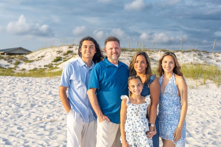 Family wearing coordinated blue and white outfits poses together on a white sandy beach at sunset